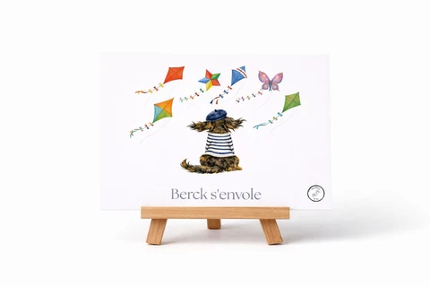 Carte illustrée d’un petit chien vu de dos, en béret bleu et haut rayé, entouré de cerfs-volants colorés, avec le titre Berck s’envole.